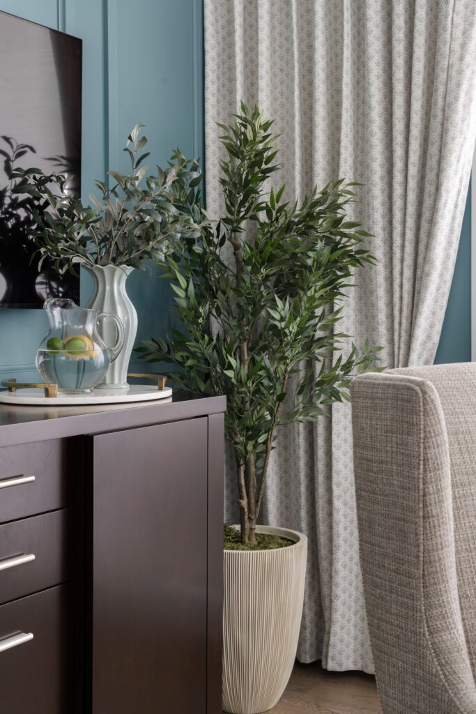 A dark wood side board cabinet holds a glass pitcher with citrus and a white ceramic vase with faux olive branches, set against a teal paneled wall beneath a mounted television. A tall faux olive tree in a fluted planter stands beside patterned curtain panels.
