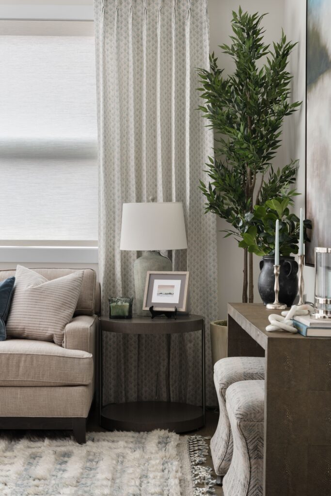 A dark wood side table holds a ceramic table lamp and a small framed photo, positioned beside a cream sofa and a tall faux olive tree in a fluted planter. A patterned drapery panel frames the window behind, with a dark console and decorative vase visible at right.
