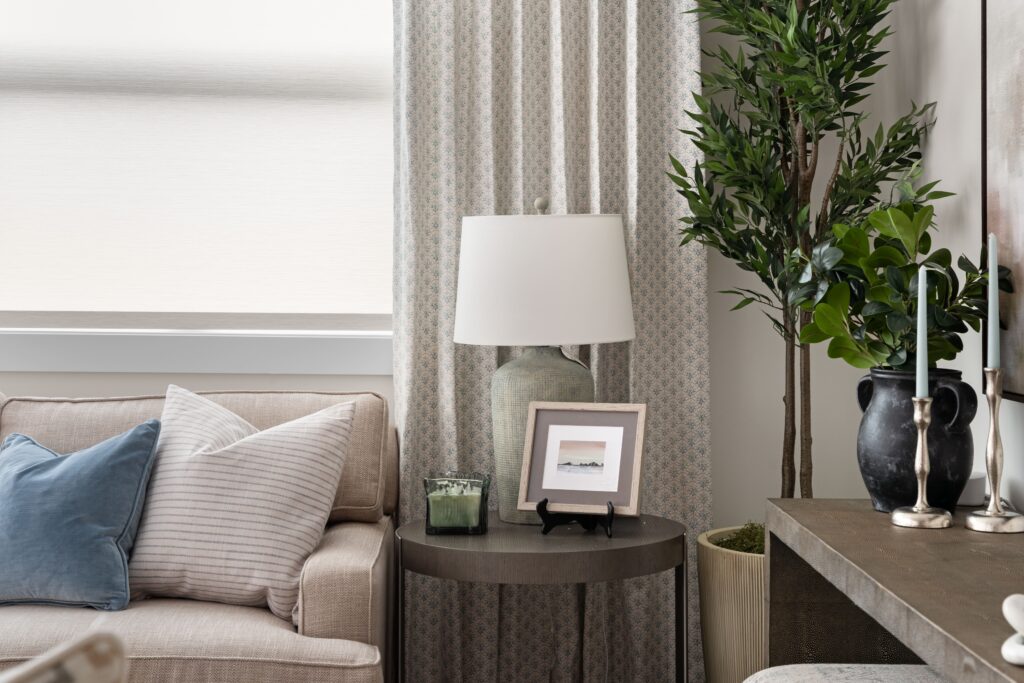 A dark round side table holds a ceramic table lamp, a small framed photo, and a candle, positioned beside a cream sofa with striped and blue pillows. A faux olive tree and silver candlesticks on a dark console are visible to the right.
