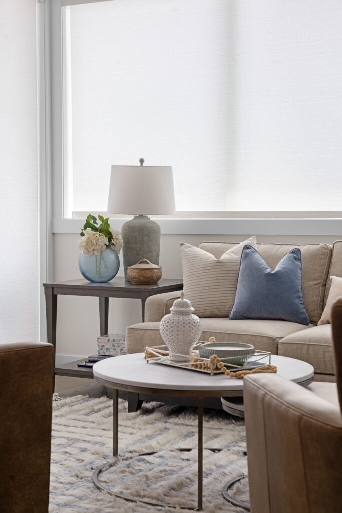 A round marble-top coffee table holds a white ginger jar, a green candle, and a set of light wood beads, set in front of a cream sofa with a blue velvet throw pillow. A dark wood side table beside the sofa holds a ceramic table lamp and a blue glass vase with white flowers.