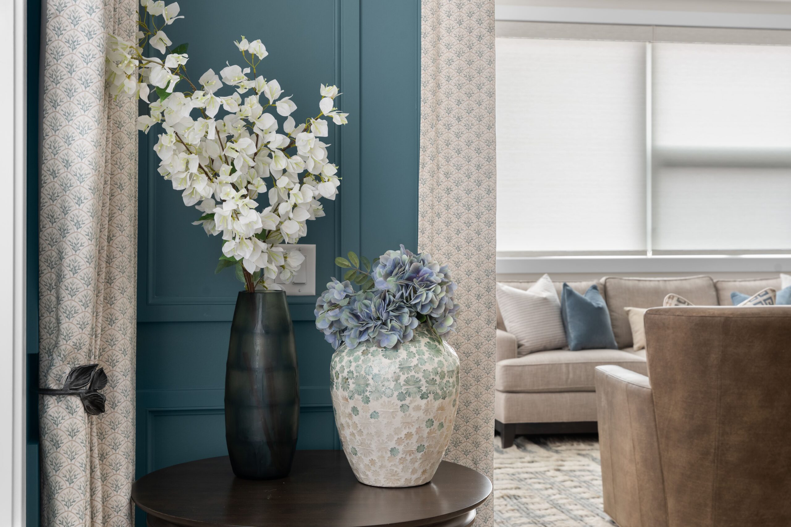 A dark wood table displays a black textured vase with white flowering branches and a mosaic-patterned vase with blue hydrangea, set within a teal paneled corner. Patterned curtain panels frame the corner on either side.