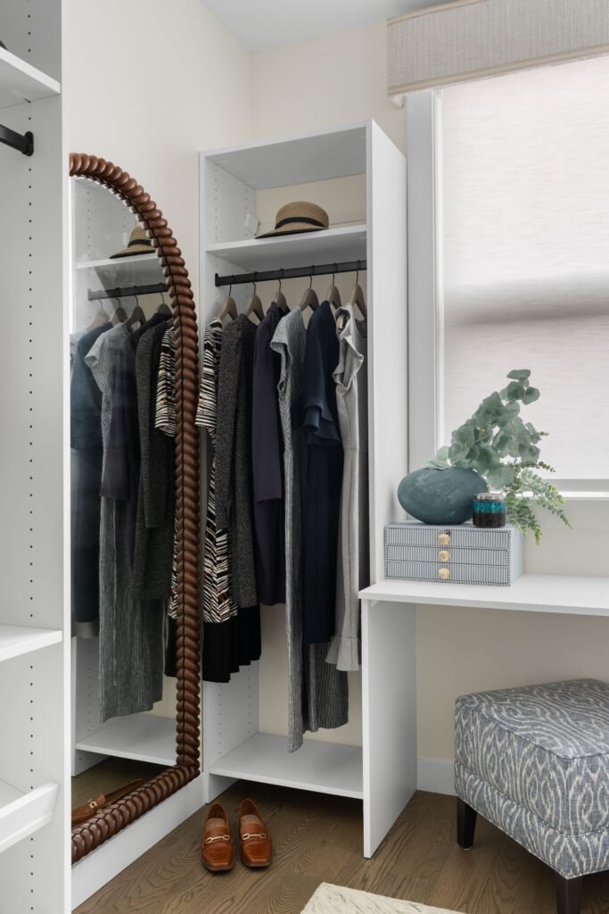 A walk-in closet features hanging rods of clothing and a full-length mirror beside a built-in vanity nook styled with a dark blue vase, faux eucalyptus, and a small storage box. A patterned upholstered ottoman and a pair of leather loafers sit near the vanity.