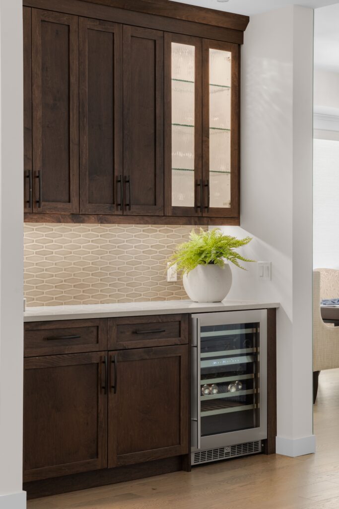 Dark wood bar cabinetry with glass-front upper doors and a built-in stainless wine fridge sits beneath a light stone countertop, styled with a white ceramic planter of greenery. A geometric tile backsplash runs along the wall above the counter.