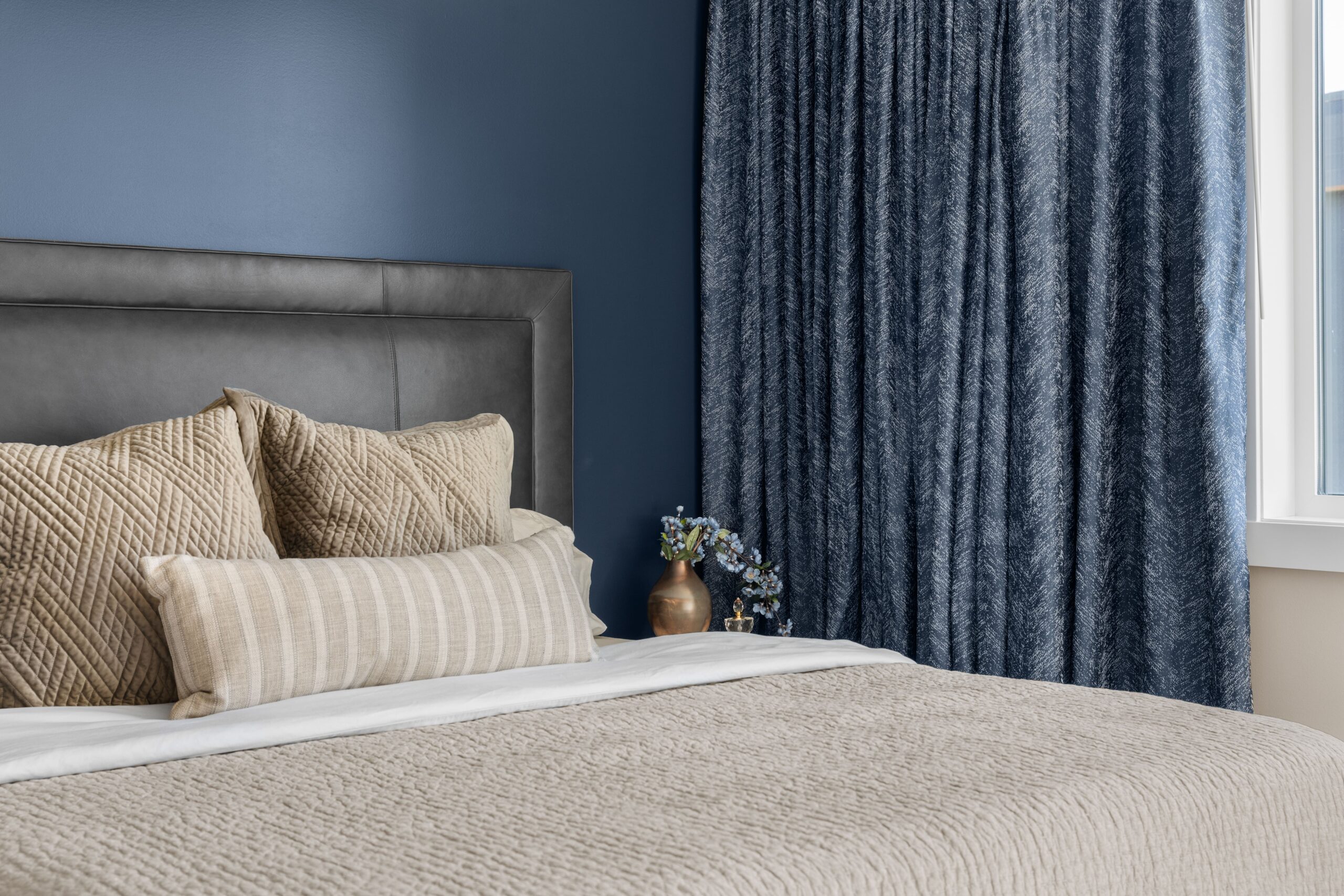 A styled bed with neutral quilted pillows and a striped lumbar pillow set against a navy accent wall and full-length blue drapery.