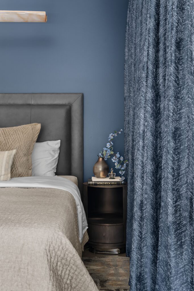 Bedroom corner pairing a dark headboard and layered neutral bedding with a round nightstand and floor-length blue curtains.