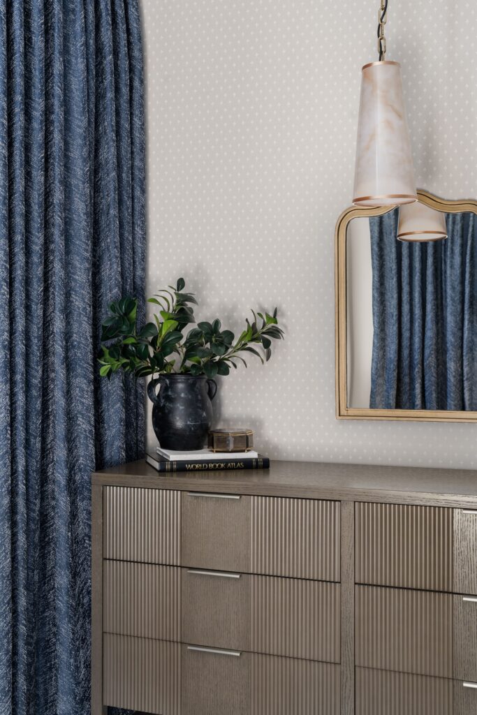 Bedroom dresser styled with greenery and books below a gold-toned arched mirror and pendant light, framed by navy drapery.