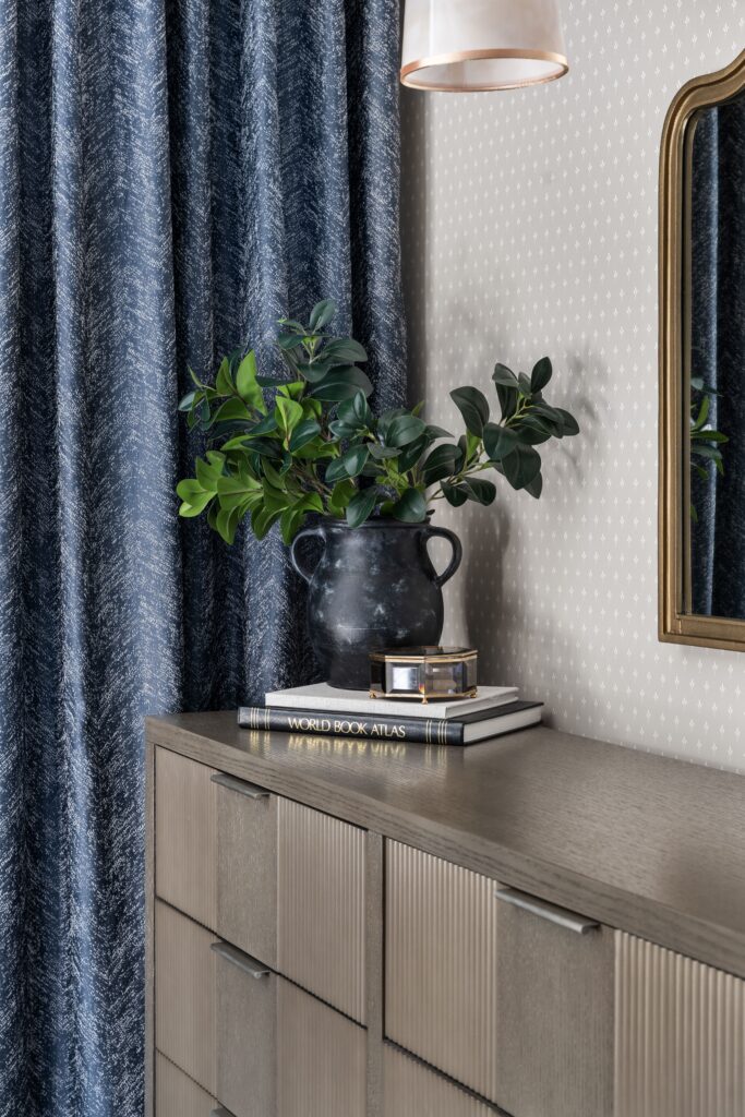Close-up of dresser styling with greenery in a black two-handled vase, stacked books and a small keepsake box beside blue curtains.