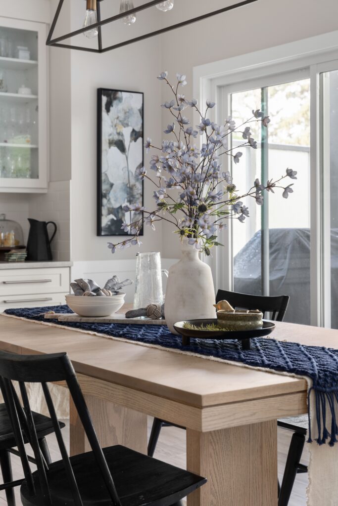 A ceramic vase and glass pitcher anchor the dining table centrepiece, surrounded by stacked bowls and folded napkins.