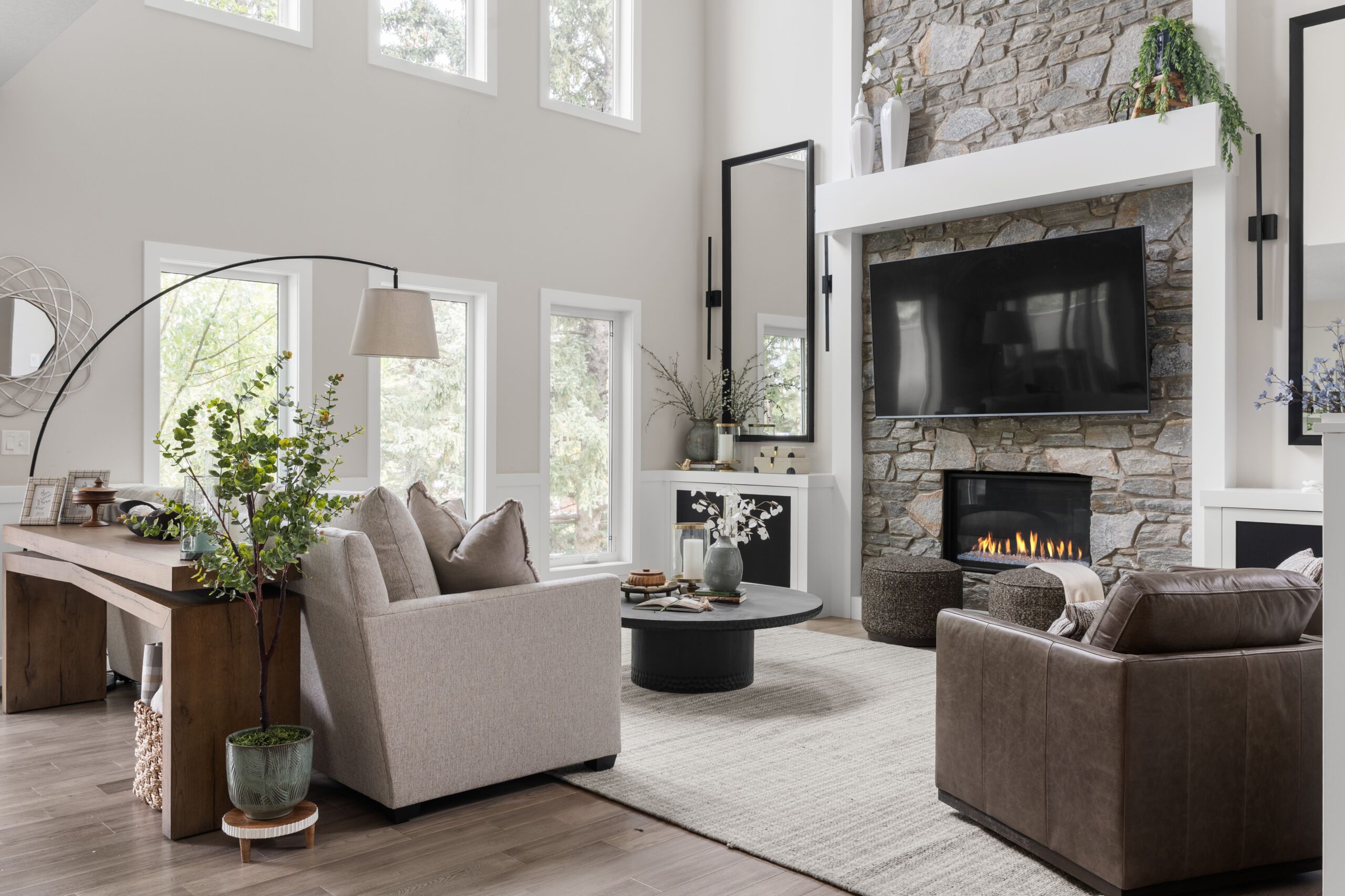 A bright, open-concept family room with a neutral sectional, swivel leather armchair, wood console table, and stone fireplace in the background.