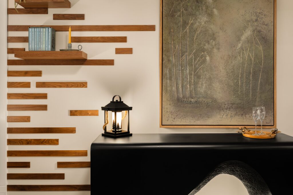 A black console table beneath a framed forest painting, flanked by a wood slat accent wall with floating shelves.