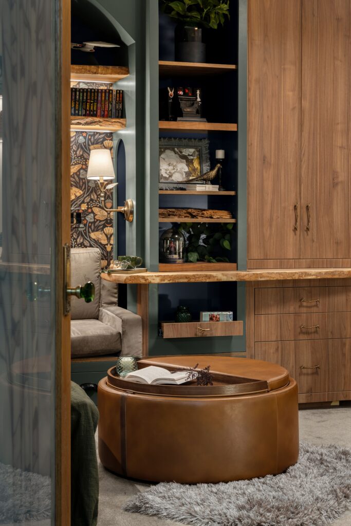 A moody built-in reading nook seen through an open glass-paned door, with a round leather ottoman and botanical wallpaper.