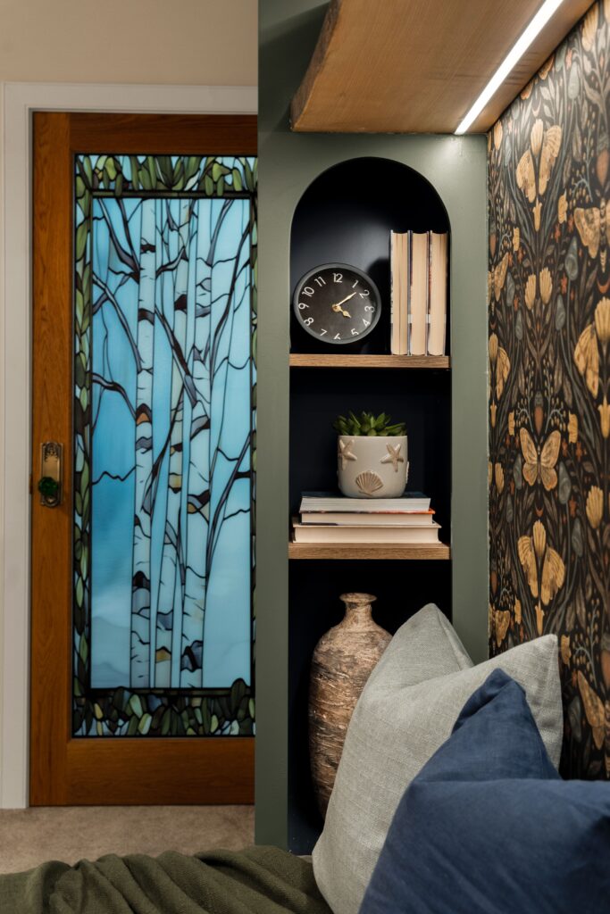 The glass-paned office door has a stained glass applique of birch trees. On the right, the botanical wallpaper is complimented by additional built-in shelving with decor and live plants.